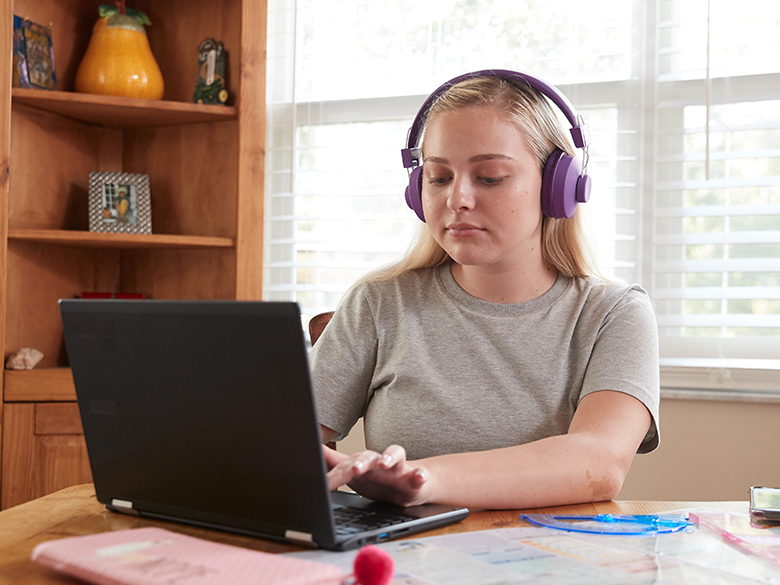 A young woman wearing purple headphones sitting at a table typing on a laptop