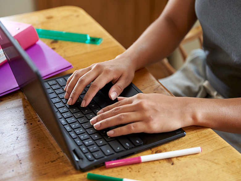 Two hands positioned on a laptop keyword on a wood table with markers, folders, and a ruler in view