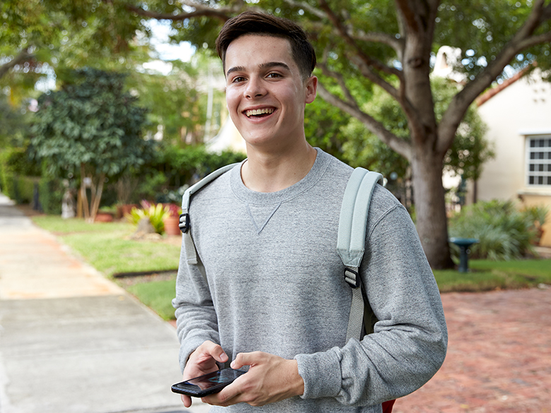 A young man smiling at the camera, standing on a sidewalk wearing a backpack while holding a smartphone