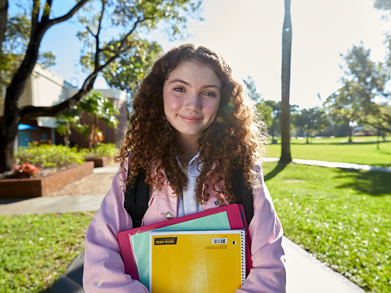 A girl smiling at the camera, standing on a sidewalk wearing a backpack while holding a notebook and binder