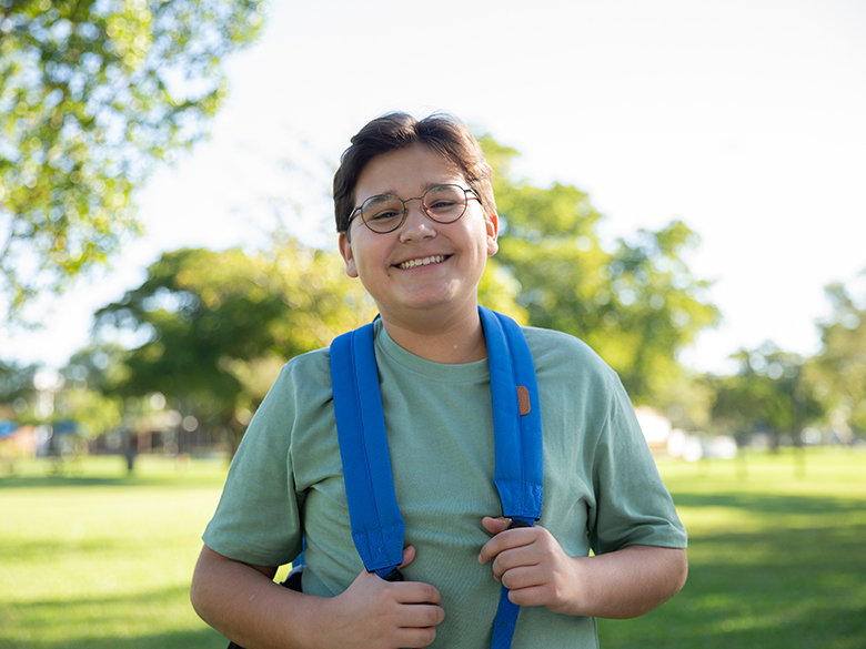 A smiling boy with glasses standing outside wearing a backpack; trees and grass in the background