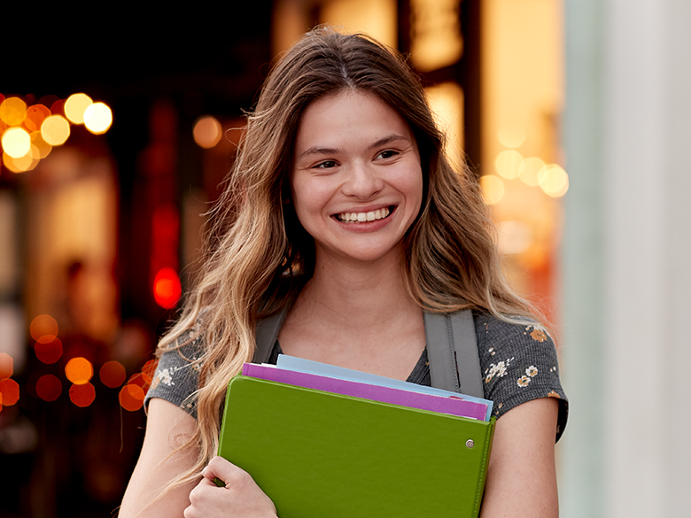 A smiling young woman wearing a backpack while holding a binder