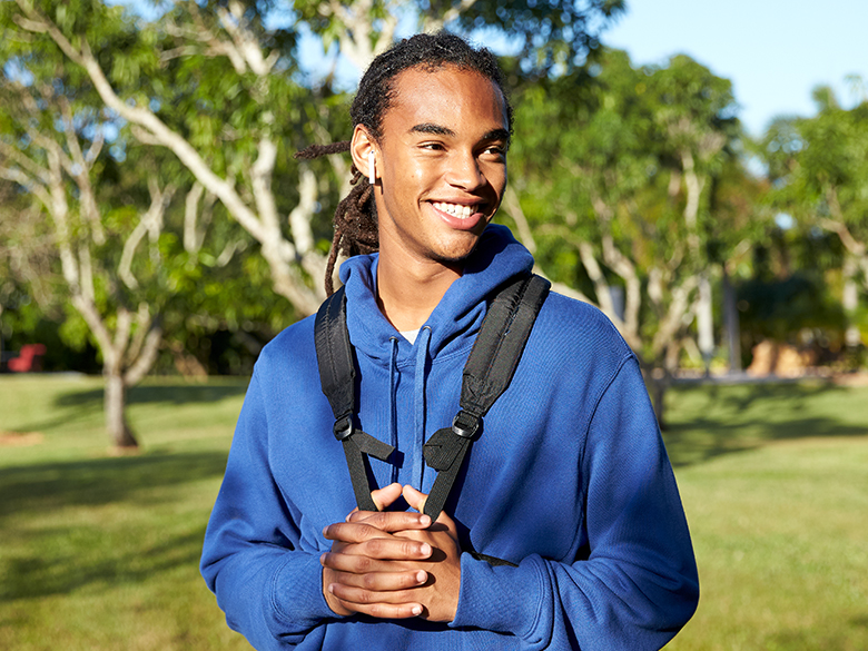 A smiling young man standing outside wearing a backpack; trees in the background