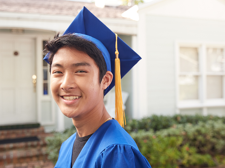 A young man standing in front of a house, smiling toward the camera, wearing a blue graduation cap and gown