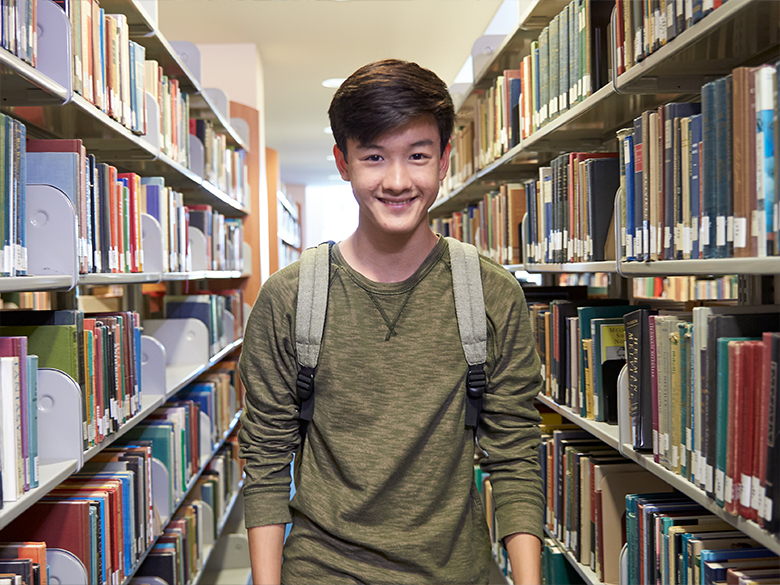 A young boy smiling toward the camera, wearing a backpack, standing in a row of bookshelves