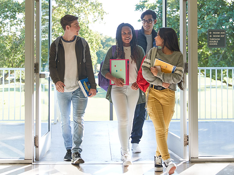 A group of four students in mid-conversation walking through open glass doors
