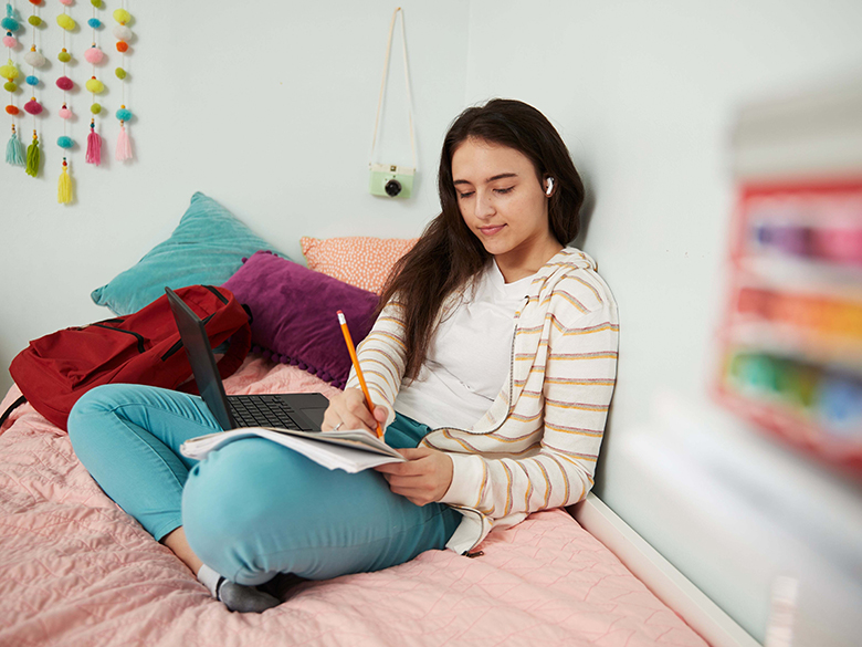 student sitting on bed, laptop computer and paper notebook in her lap