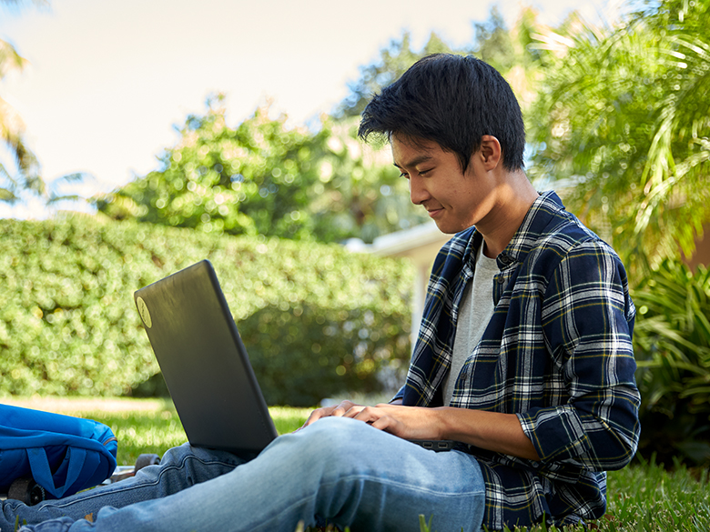 A young man sitting outside on grass typing on a laptop; trees and hedges in the background